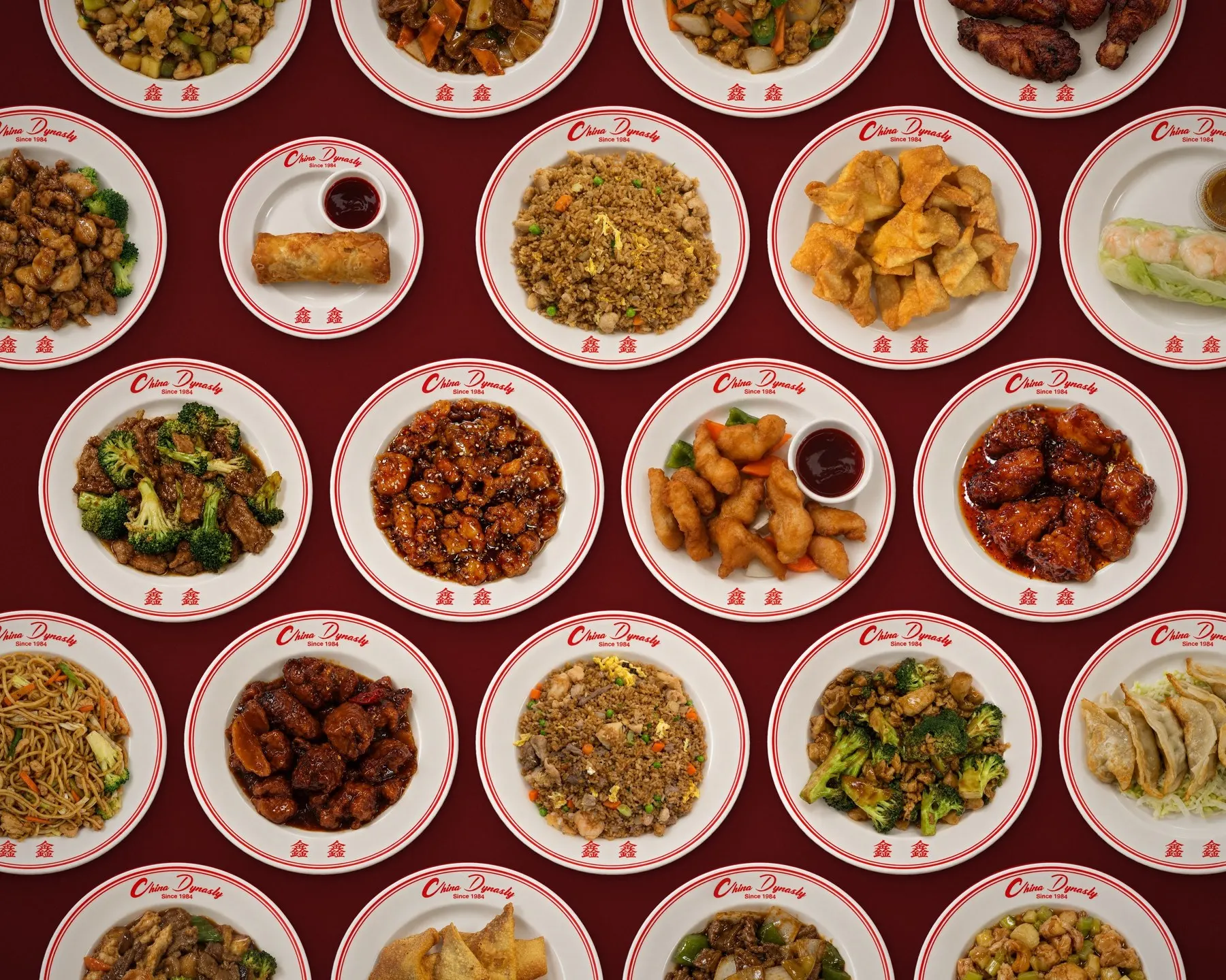 A table full of China Dynasty dishes on branded plates on a deep red table: beef with broccoli, sesame chicken, sweet and sour chicken, General Tso’s chicken, fried rice, lo mein, egg rolls, cream cheese puffs and dumplings